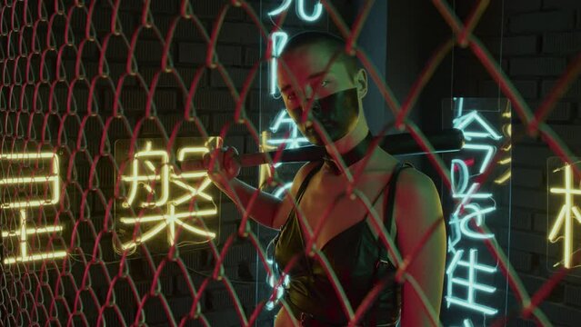 Waist-up of girl with shaved head, black paint on face, in white contact lenses hitting fencing with bat in studio with neon lights in shapes of hieroglyphs meaning nirvana and western paradise