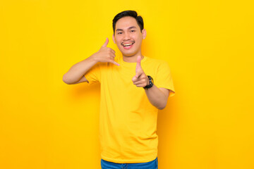 Cheerful young Asian man in casual t-shirt showing call me sign gesture and pointing at camera...