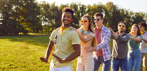 Happy multiracial friends having fun on warm sunny evening in park. Diverse group of cheerful joyful young people enjoying outdoor summer party, dancing in conga line all together and smiling - obrazy, fototapety, plakaty