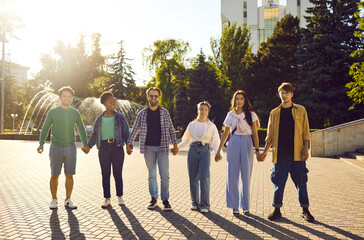 Cheerful positive multiracial friends holding hands standing in row on city street. Six joyful...