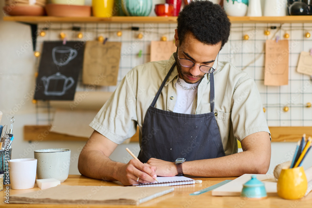 Poster African American guy in apron making notes in notepad while sitting by table and working over new collection of earthenware or clay items