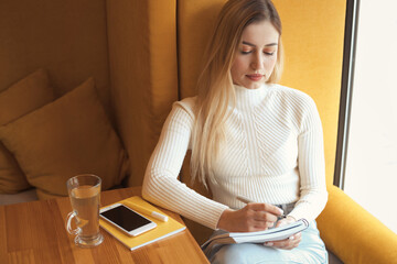 Young girl student is preparing for exam at the university and writes a task in a notebook in cafe