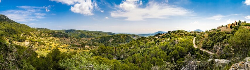 Panorama of the valley of the mountain village Galilea at Mallorca in the municipality of Puigpunyent surrounded by mountains, mediterranean forest and olive groves with view to the mediterranean sea. - obrazy, fototapety, plakaty