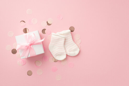 Baby concept. Top view photo of white gift box with ribbon bow tiny socks and shiny confetti on isolated pastel pink background