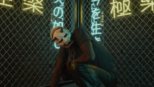 Tracking in of young Black man with white hair in mask of demon sitting in studio with netting fence and neon lights meaning nirvana and western paradise