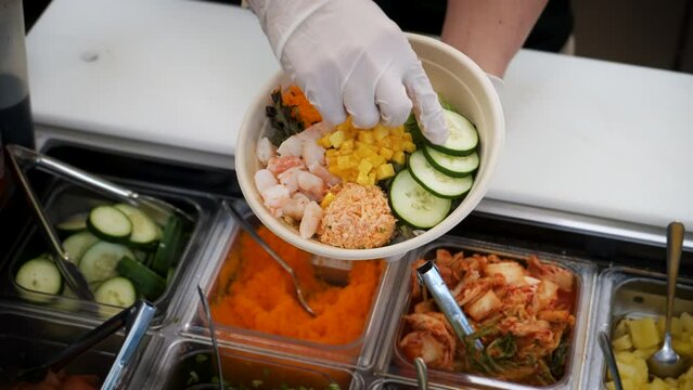 Top down view of worker building a traditional sushi salad, adding cucumber to poke bowl from assembly station, slow motion 4K