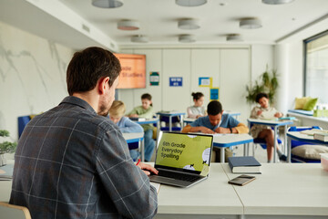 Back view at male teacher using laptop at desk in classroom with group of children studying in...