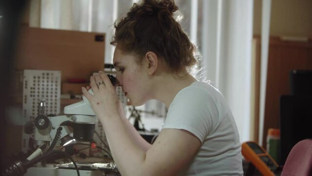 Female worker at a service center using microscope and to electronics circiuit board. Girl looking through a microscope in workshop. Women and technical occupations.