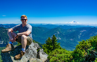Naklejka premium An athletic adventurous male hiker sitting on top of a mountain looking at the camera smiling with a beautiful mountain range on a sunny day in the background.