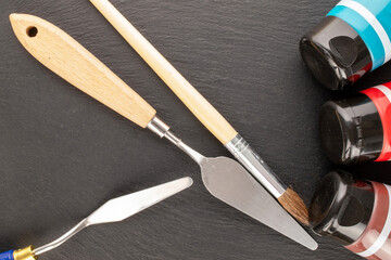 Two metal palette knives, a brush and paints on a slate stone, close-up, top view.