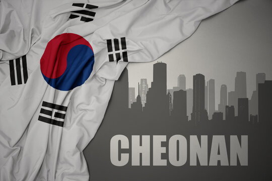 abstract silhouette of the city with text cheonan near waving national flag of south korea on a gray background.