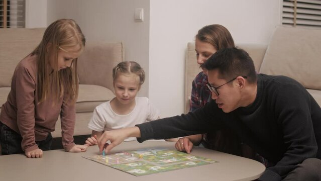 Medium shot of multi ethnic family of different age generations are playing a board game, throwing dice on the table at home sitting on the floor. Mom dad and their daughters play family games in the