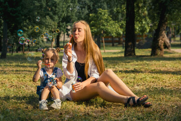 Fototapeta premium Mom and daughter have fun blowing soap bubbles sitting on the grass in nature in the park