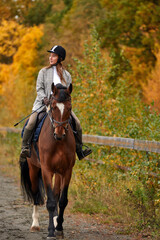 Young beautiful brunette girl rides a horse on a warm and sunny autumn day. Portrait of a pretty young woman on the horse, wearing tall boots and gloves.