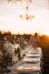 The delicate decor of the wedding banquet table is in pastel colors. Wildflowers, crystal glassware, candles. Сrystal chandelier on top, illuminating the table. Rays of the setting sun.