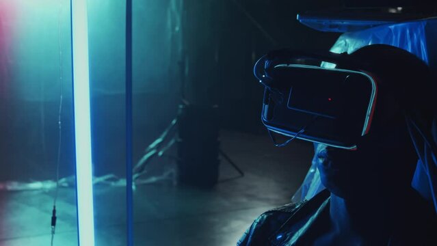 Shoulder-up of young Black guy in virtual reality goggles sitting in hideout with plastic film on walls and lights twitching