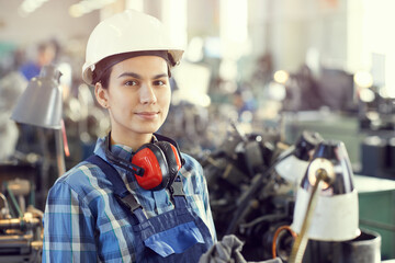 Portrait of content attractive female manual lathe worker in work helmet and ear protectors around...
