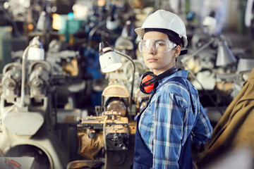 Portrait of serious tomboy in hardhat and protective goggles working with lathe at factory