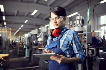 Busy young factory woman in blue checkered shirt and overall standing in industrial shop with...