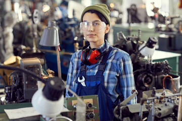 Portrait of serious hipster girl in hat and protective eyewear standing at manual lathe and repairing it
