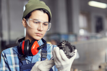 Serious thoughtful hipster girl in protective goggles standing in factory shop and watching milling machine tool