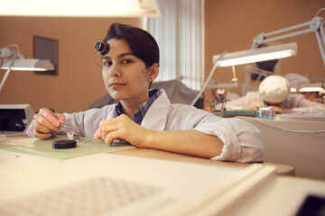 Portrait of serious confident young woman in white coat sitting at desk and assembling mechanical...