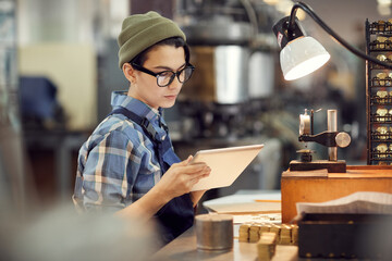 Serious busy hipster female worker of watch factory sitting at desk and reading online instruction...