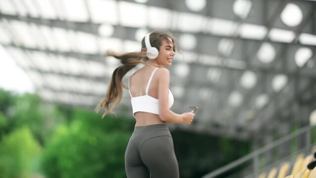 Smiling athletic young woman in fashion sportswear holding smartphone and enjoying favorite song at stadium. Workout Playlist. A beautiful girl listens to music and dances in between sports