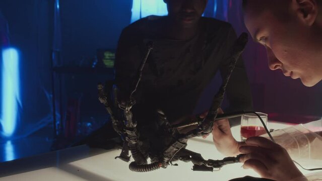 Over shoulder slowmo of Caucasian cyberpunk girl with shaved head soldering robotic arm of Black man who sitting at table in front of her in dark room with neon lights, drinking