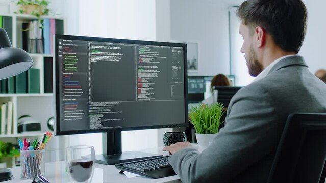 Dolly out Over the Shoulder Shot of Young Businessman Sitting at Desk Using Desktop Computer Writing Program Code. Bright Office with coworkers. Caucasian Male Programmer or Software Engineer.
