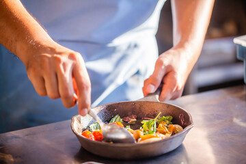 Chef cooking food in restaurant kitchen. Gourmet chef in uniform preparing meal, close up. Male in apron stirs with a spoon vegetables in frying pan on kitchen counter in luxury fine dining restaurant
