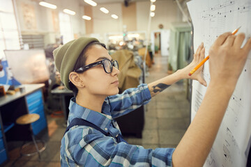 Side view of focused young woman in glasses standing at factory shop and drafting technical sketch...