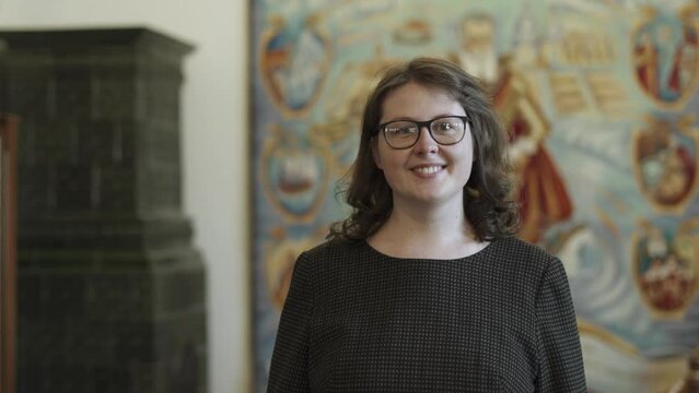 Young pretty woman with glasses and curly hair smiles, looks at camera and around in historical hall of the castle, royal library. Portrait of a happy, satisfied researcher, librarian, teacher.