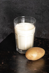 Glass of potato milk and potato tuber on old rusty metal surface on black backdrop. Selective focus.
