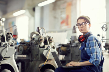 Serious smart hipster girl in eyeglasses sitting at factory and using laptop while working with...