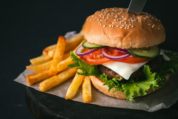 Big beef patty burger with cheese tomato purple onion cucumber and lettuce next to french fries on wooden black stand on black background