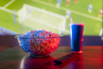 Football match on the TV screen. Popcorn in a bowl, a drink in a glass and a remote control. Cozy...