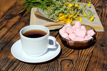 On a wooden table is a cup of tea with a book and a bouquet of wildflowers.