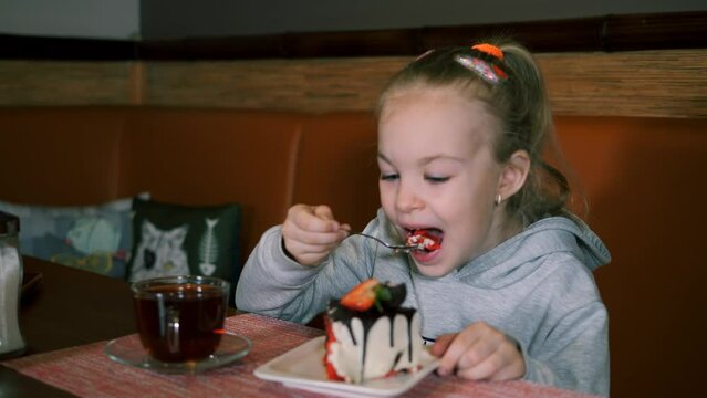Little girl eats dessert with fresh strawberries and drinks tea. Child smiles sweetly while eating. The baby brings a fresh strawberry to her mouth.