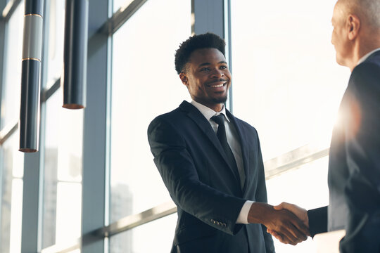 Cheerful handsome young Black businessman in suit standing at panoramic window and making handshake with business partner in lobby of company