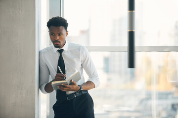 Fototapeta premium Serious pensive young Afro-American employee leaning on wall in hall and making notes in organizer while planning work day