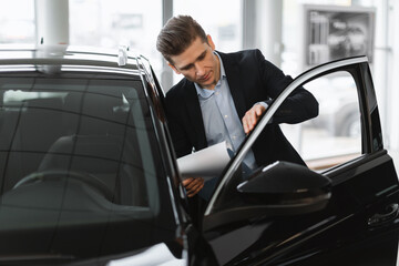 Serious young auto insurance agent checking new car, taking notes, working at automobile dealership