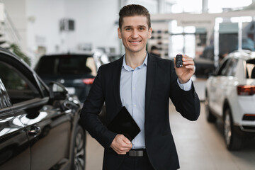 Handsome young car salesman showing car key at camera, selling autos at dealership