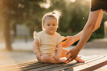 A female toddler in yellow clothes is enjoying the summer on the bench near the hands of a mommy in the park in the sun rays. Mother is protecting her young daughter from falling at sunset.