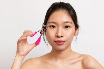 Closeup portrait of pretty asian lady using eye lash curler on long eyelashes, posing on white studio background