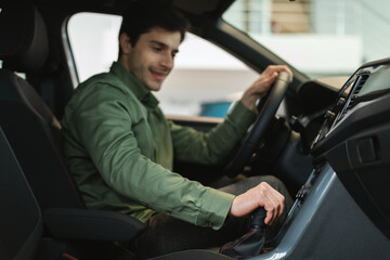 Fototapeta na wymiar Cheerful young Caucasian guy test driving new car before purchase at automobile dealership, selective focus