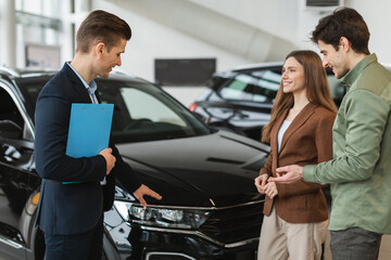 Positive young spouses speaking to salesman about purchasing new auto at dealership shop. Car...