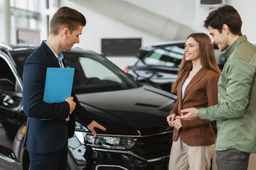 Fototapeta premium Positive young spouses speaking to salesman about purchasing new auto at dealership shop. Car purchase or rental