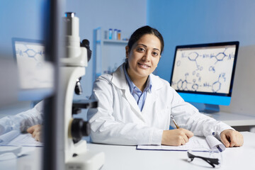 Portrait of smiling young Indian woman in lab coat sitting at desk and making notes in clipboard...