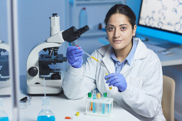 Portrait of content young Indian female pharmacist in lab coat and surgical gloves sitting at desk...